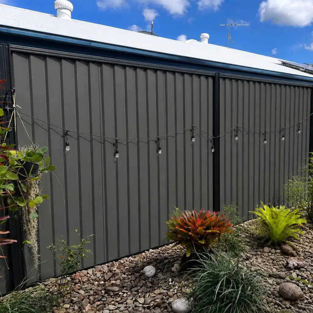 Gray corrugated metal fence with a string of outdoor lights surrounded by rocks and desert plants under a blue sky