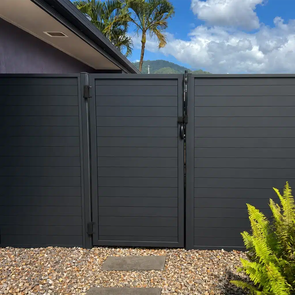 Dark gray metal gate with horizontal slats and a central latch set in a fenced enclosure over a gravel ground with tropical plants and blue sky in the background