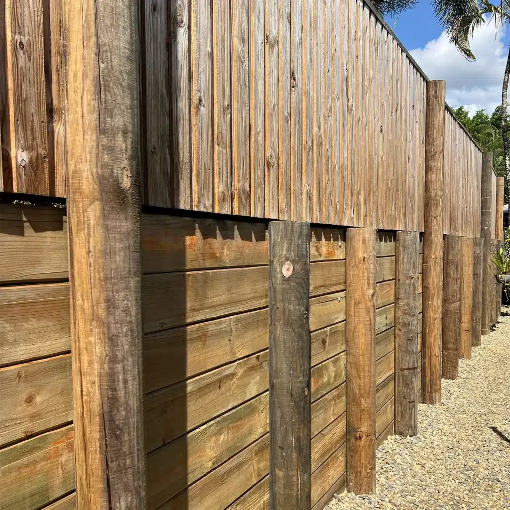 Tall weathered wooden fence with vertical boards and sturdy posts along a gravel path under a blue sky with palm trees in the background