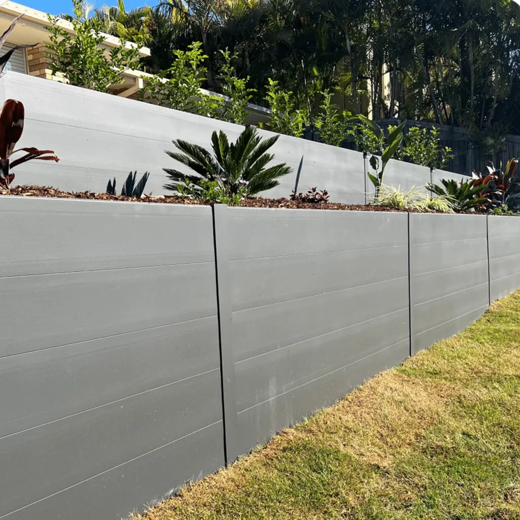 Gray multipanel privacy fence on a sloped yard with tiered planter beds and tropical plants along the top edge blue sky visible above